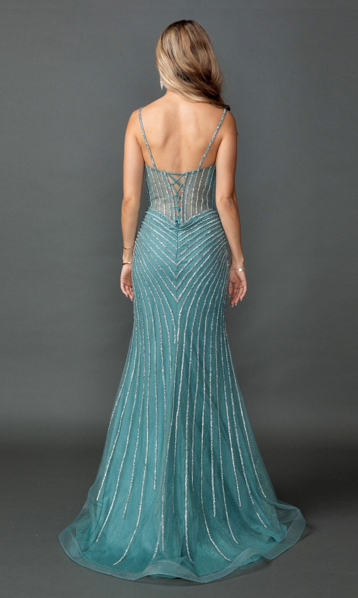 A woman with long hair stands facing away, wearing the Dancing Queen 4574—a sleeveless, teal, floor-length prom dress. This gown features thin straps and silver beaded lines arranged in an elegant chevron pattern, creating a stunning beaded prom look.
